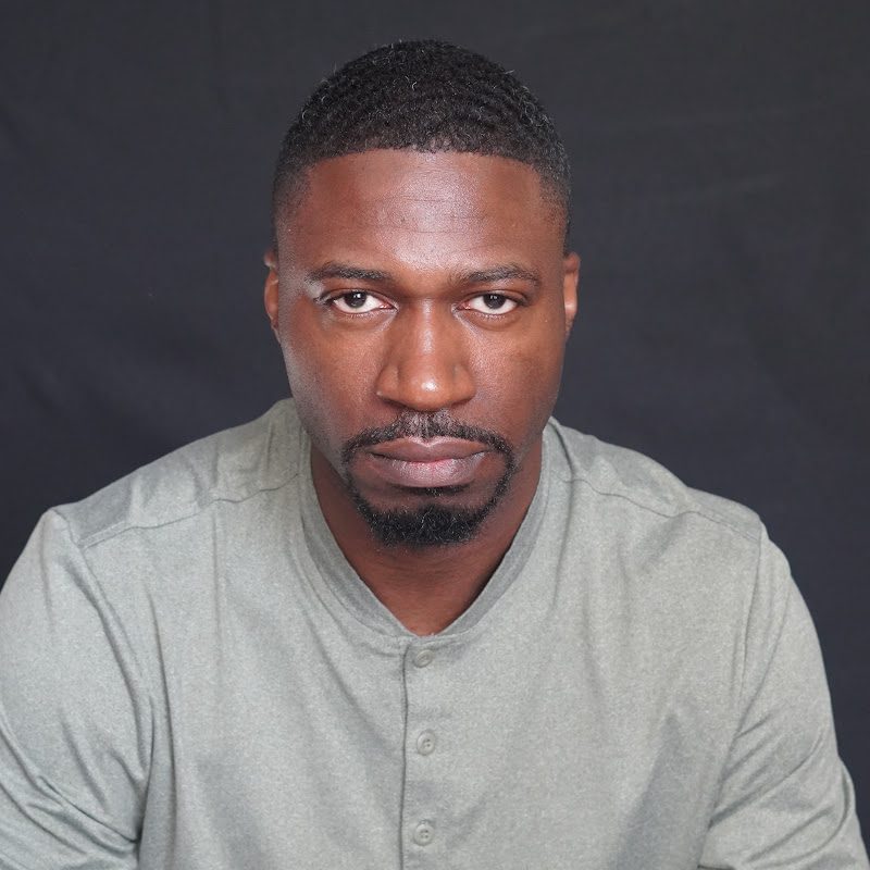 Portrait of NovA in a grey shirt against a dark backdrop, looking directly at the camera
